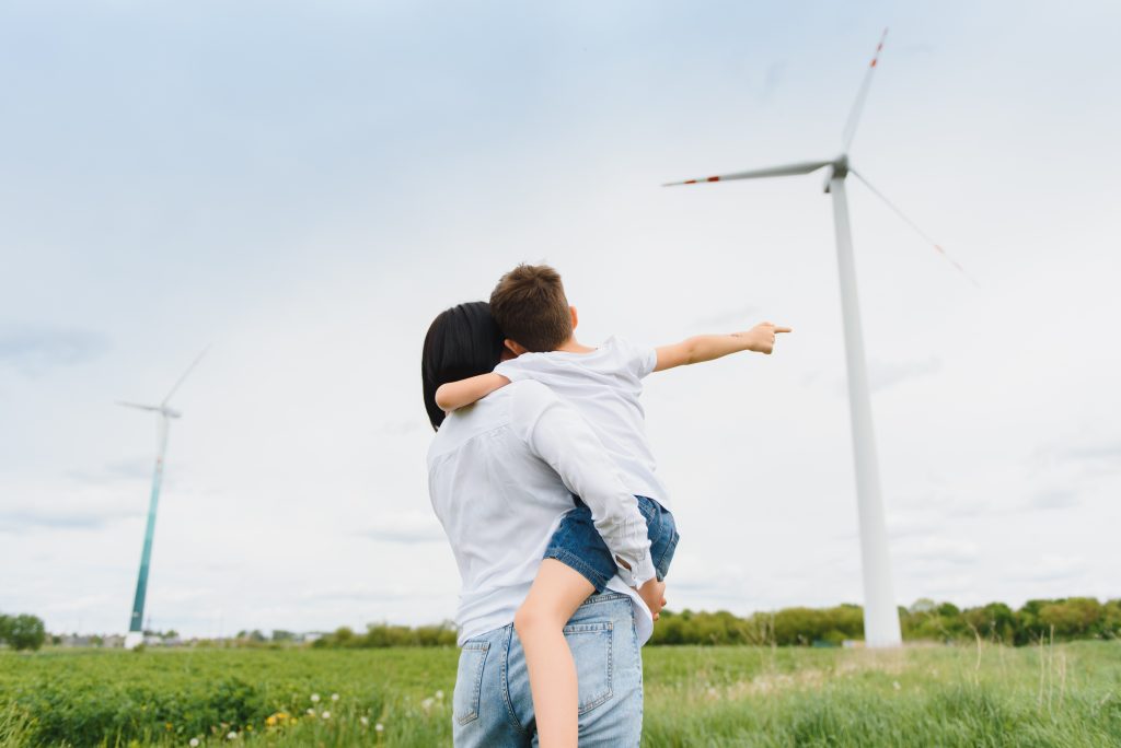 A mom holding her son in a field pointing at a windmill while she tells him about the future of her investments.